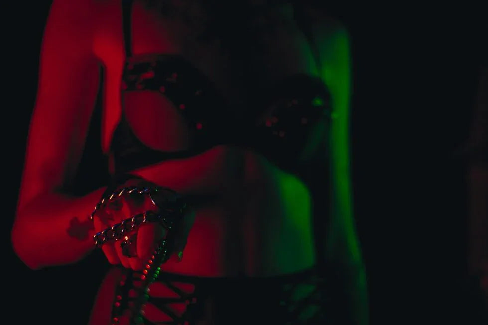 Woman in black studded lingerie holding metal chain in dim red and green lighting BDSM FemDom style