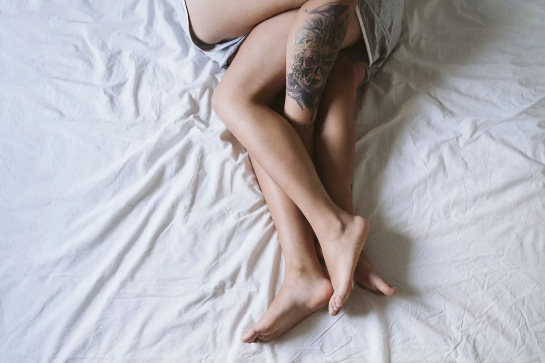 Two pairs of intertwined bare legs on white bed sheets, one leg with a detailed tattoo
