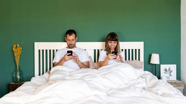 Couple in white bedding using smartphones in bed against green wall, modern bedroom setting