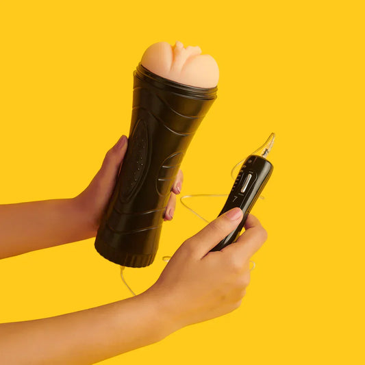 Hands holding a black male masturbator with a flesh-colored sleeve and wired remote control on yellow background