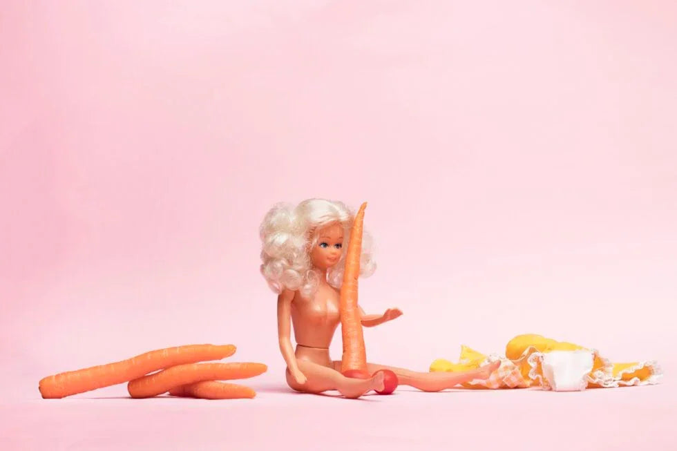 Naked doll with curly blonde hair sitting on pink surface holding a large carrot, surrounded by multiple carrots and doll clothes